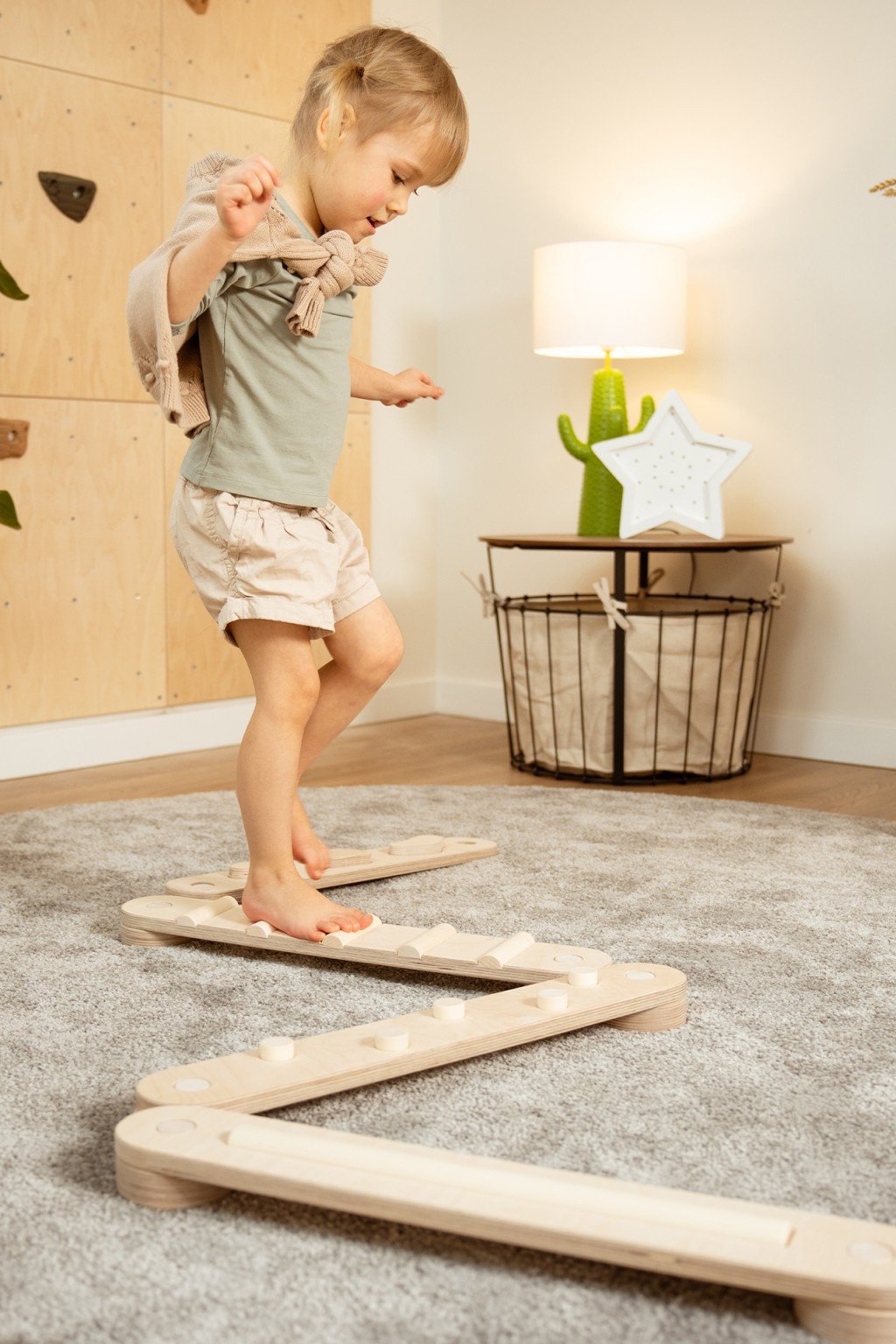 Enfant en bas âge marchant sur un chemin en bois posé sur tapis, intérieur chaleureux avec lampe et panier décoratif. Mettre un bébé dans un parc n’est pas ici.