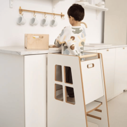 Enfant debout dans une cuisine Montessori, utilisant une tour d'observation blanche avec marches et ouverture en bois. hauteur des tours d'observation adaptée.