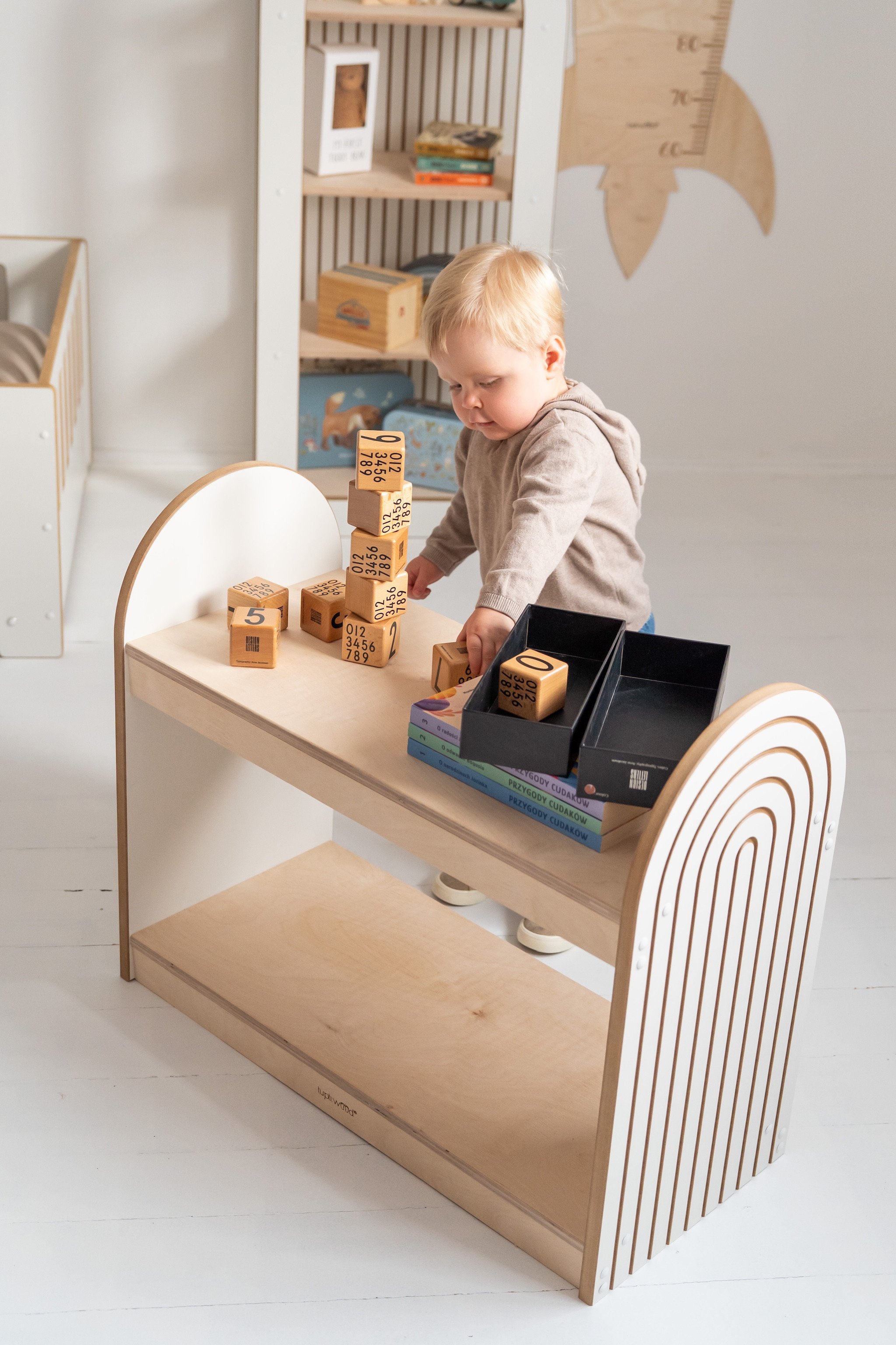 Enfant jouant avec des cubes en bois sur une table basse, rangement livres enfant accessible et étagères en arrière-plan dans une pièce lumineuse.