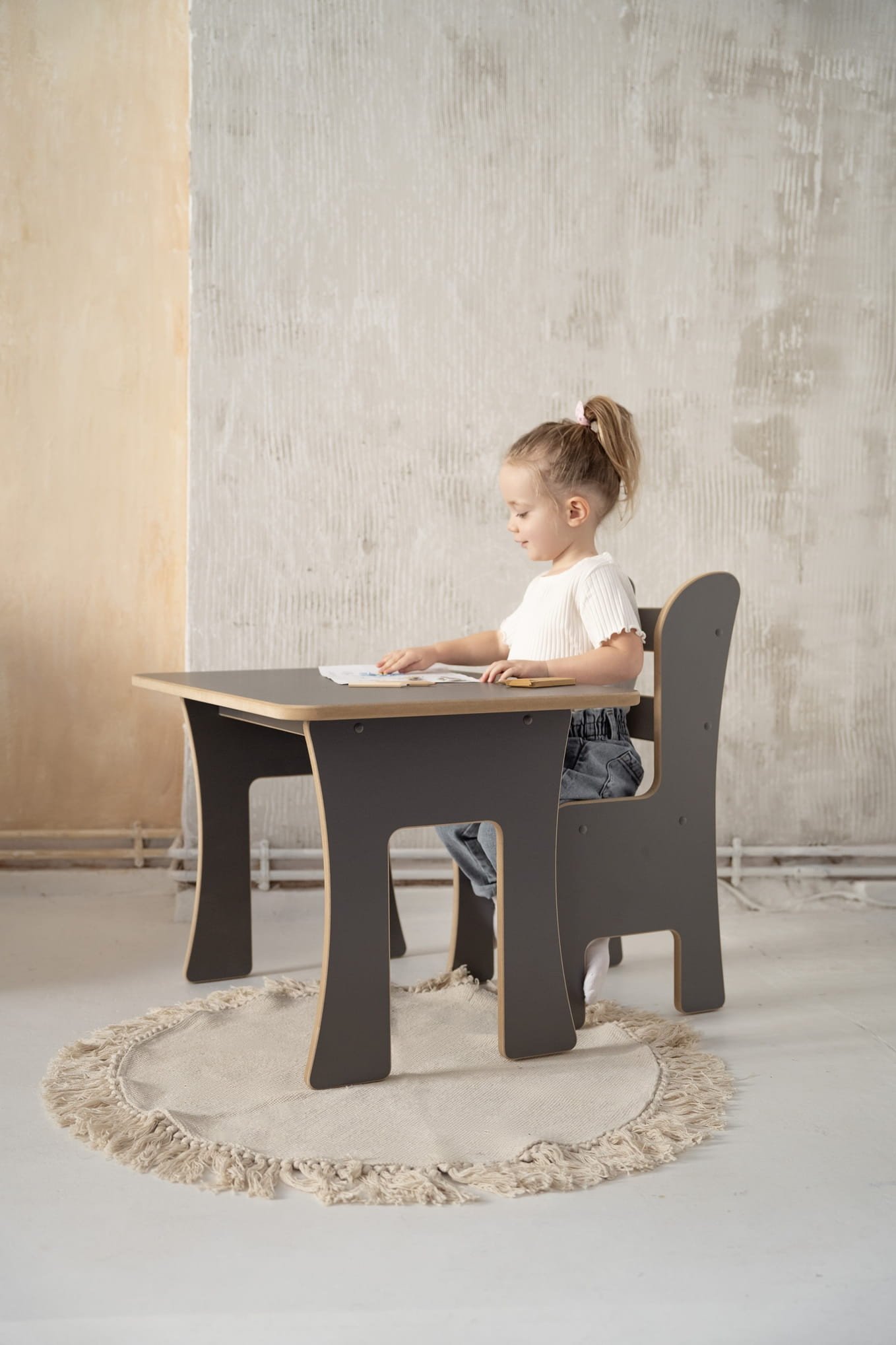 Petite fille assise à un petit bureau, concentrée sur une activité, dans une pièce au mur brut, sur un tapis rond clair. intégration du mot-clé: comment développer l'autonomie d'un enfant.