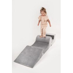 Petite fille marchant sur un module pliable gris, part de parcours d’agilité pour jeu intérieur. Piscine à balles enfant montessori non visible mais suggérée par l’idée d’éveil et motricité.